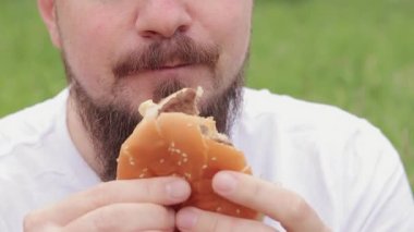 Yakın çekim erkek yüzü büyük bir burger yiyor. Sakallı aç genç adam lezzetli bir burger yiyor. Adamın ağzı büyük bir burgeri yakından ısırıyor. Doğada atıştırmalık