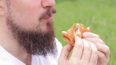Yakın çekim erkek yüzü büyük bir burger yiyor. Sakallı aç genç adam lezzetli bir burger yiyor. Adamın ağzı büyük bir burgeri yakından ısırıyor. Doğada atıştırmalık