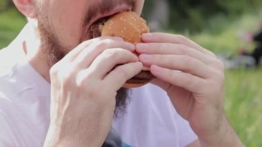 Yakın çekim erkek yüzü büyük bir burger yiyor. Sakallı aç genç adam lezzetli bir burger yiyor. Adamın ağzı büyük bir burgeri yakından ısırıyor. Doğada atıştırmalık