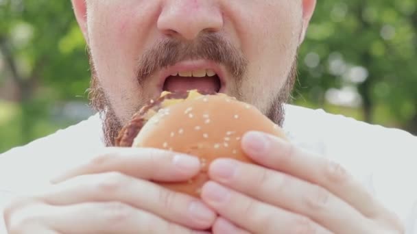 密切的男性脸吃一个大汉堡 饥饿的留着胡子的年轻人吃着美味的汉堡包 那家伙的嘴咬一个大汉堡特写 小菜一碟 — 图库视频影像