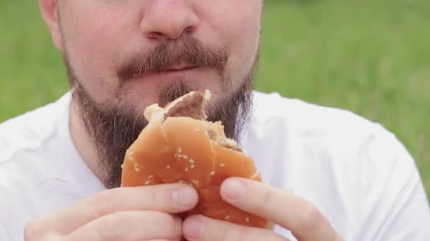 密切的男性脸吃一个大汉堡 饥饿的留着胡子的年轻人吃着美味的汉堡包 那家伙的嘴咬一个大汉堡特写 小菜一碟 — 图库视频影像