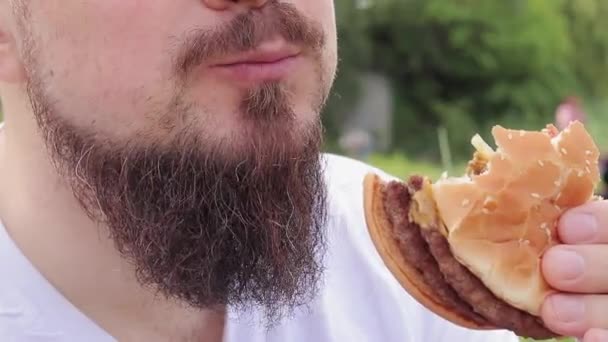 ハンバーガーを食べる クローズアップ男性の顔はチーズバーガーを食べています 髭を生やした空腹の青年がおいしいバーガーを食べる 男の口は食べ物を噛んでいる 自然の中で軽食 — ストック動画
