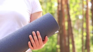 Elinde spor malzemesi ve spor paspası tutan aktif bir kız. Ormanda açık havada spor. Kız antrenmanına dışarıda başlıyor. Elinde spor minderi ve yoga olan bir kız, yakın plan.
