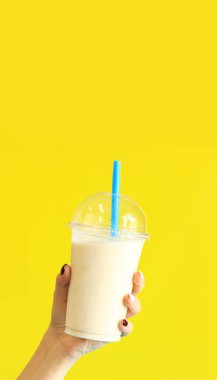 Plastik bardaklı muzlu smoothie. Muzlu milkshake, parlak sarı arka planda bir kızın elinde. Boşluğu kopyala