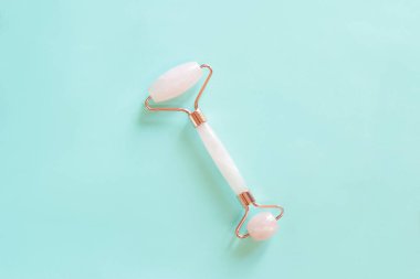 Mavi arka planda gül kuvars yüz masajı silindiri. Yüz masajı aleti, kırışıklık önleyici cilt bakımı aracı, yazı için yer. Doğal kozmetik ürünleri ve pastel arka planda yüz bakımı ürünleri, üst görünüm