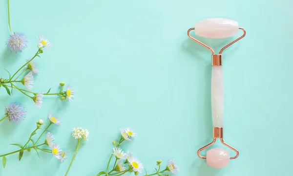 stock image Facial massage roller made of rose quartz on a blue background with flowers. Facial massager anti-wrinkle anti-aging skin care tool. Natural cosmetics and face care products on a pastel background