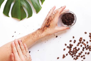 Kadın elleri cildine kahve fırçası sürerler. Doğal kozmetik konsepti. Kahve fırçası, el, kahve çekirdeği ve canavar yaprağından oluşan düz bir kompozisyon. Beyaz arkaplan üzerindeki spa prosedürü