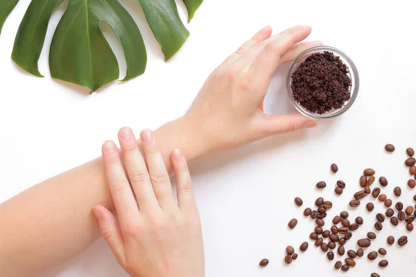 Women\'s hands and coffee scrub on the skin. The concept of natural cosmetics. Flat lay composition of coffee scrub, hands, coffee beans and monstera leaf. Spa procedure on a white background