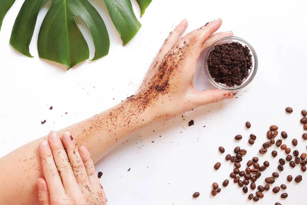 Female hands apply coffee scrub to the skin. The concept of natural cosmetics. Flat lay composition of coffee scrub, hands, coffee beans and monstera leaf. Spa procedure on a white background