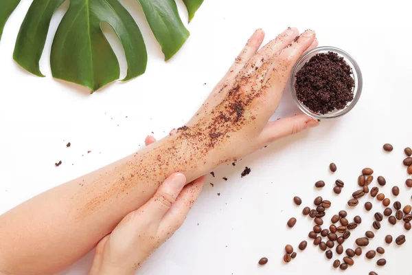 Female hands apply coffee scrub to the skin. The concept of natural cosmetics. Flat lay composition of coffee scrub, hands, coffee beans and monstera leaf. Spa procedure on a white background