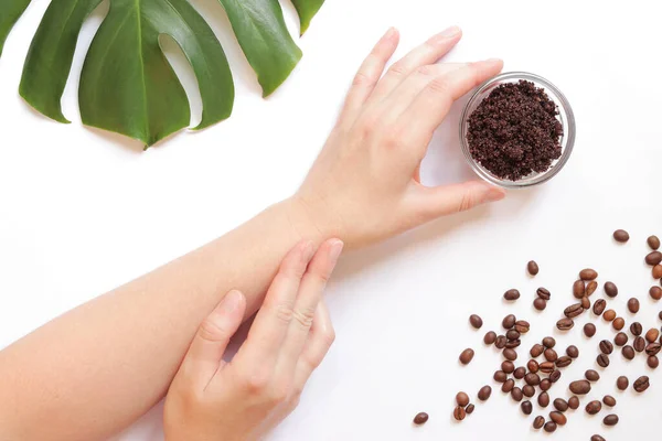 Women\'s hands and coffee scrub on the skin. The concept of natural cosmetics. Flat lay composition of coffee scrub, hands, coffee beans and monstera leaf. Spa procedure on a white background