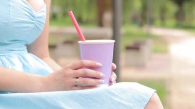 Mavi elbiseli bir kız parkta limonata içiyor. Yakından çekilmiş bir kadının ellerinde bir fincan lavanta rengi. Bir içki al. Tazeleyici içecek.