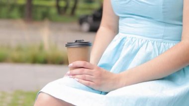 Mavi elbiseli bir kız parkta kahve içer. Bir kadının ellerinde kahve olan el işi bardaklar. Hazır kahveyi al. Canlandırıcı bir içecek.