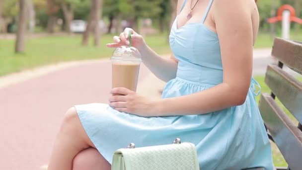 青いドレスの女の子が公園でコーヒーを飲んでいる 女性の手のひらにコーヒーが入ったプラスチックカップ テイクアウトコーヒー 飲み物を活性化する アイスラテ — ストック動画