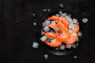 Koyu ahşap zemin üzerinde buzla kaplı bir tabakta büyük soğutulmuş karides. Siyah bir tabakta buzlu haşlanmış karides. Siyah arka planda taze karides, üst manzara. Ezilmiş buz üzerinde deniz ürünleri.