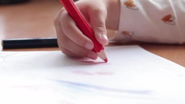 Bir anaokulu çocuğu bir albümde renkli kalemlerle resim çizer. Kalemle yakın plan, seçici odaklı çocuk eli. Kız kırmızıyla kalpler çiziyor.