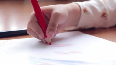 Bir anaokulu çocuğu bir albümde renkli kalemlerle resim çizer. Kalemle yakın plan, seçici odaklı çocuk eli. Kız kırmızıyla kalpler çiziyor.