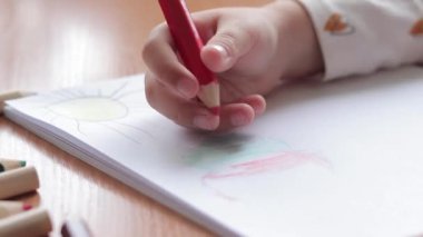 Bir anaokulu çocuğu bir albümde renkli kalemlerle resim çizer. Kalemle yakın plan, seçici odaklı çocuk eli. Kız renkli bir bulut çiziyor.