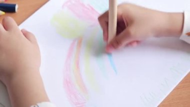 Bir anaokulu öğrencisi bir albümde renkli kalemlerle resim çizer. Kalemle yakın plan, seçici odaklı çocuk eli. Kız gökkuşağı çiziyor