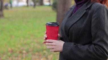 Sonbaharda bir kız parkta kahve ya da çay içer. Elleri kağıt bardakla yakın plan. Soğuk havada sıcak içecekler için. Bir fincan kahveli kadın elleri.