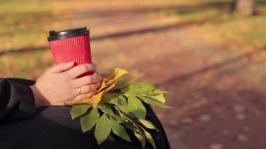 Sonbaharda kız parkta kahve ya da çay içer. Elleri kağıt bardakla yakın plan. Soğuk havada sıcak içecekler için. Bir fincan kahve ve sonbahar yapraklı kadın elleri.