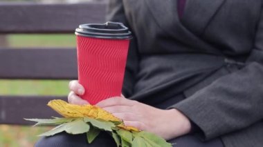 Sonbaharda kız parkta kahve ya da çay içer. Elleri kağıt bardakla yakın plan. Soğuk havada sıcak içecekler için. Bir fincan kahve ve sonbahar yapraklı kadın elleri.