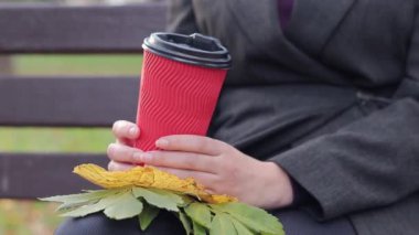 Sonbaharda kız parkta kahve ya da çay içer. Elleri kağıt bardakla yakın plan. Soğuk havada sıcak içecekler için. Bir fincan kahve ve sonbahar yapraklı kadın elleri.
