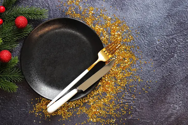 Black plate, fork and knife, gold glitter, top view of Christmas composition. Dishes and fir branch with decor on a gray table. New Year. Christmas dinner, cutlery