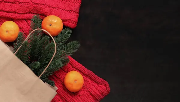 A spruce branch in a craft bag, a red scarf and ripe tangerines, top view. Christmas composition, New Year. Buy a small Christmas tree or twig for the holiday. Copy space