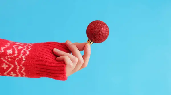A woman's hand in a red New Year's sweater holds a Christmas ball, blue background. Christmas decor, red Christmas tree toy close-up. Banner, copy space
