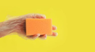 Sarı arka planda bir kadının elinde turuncu bulaşık süngeri. Şeffaf tek kullanımlık eldivendeki bir el ıslak temizleme için bir sünger tutar. Profesyonel temizlik. Boşluğu kopyala