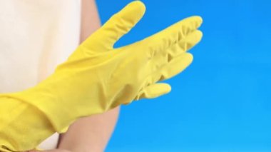 Odayı temizlemek ve bulaşıkları yıkamak için lateks eldivenli kadın elleri. Profesyonel temizlik. Ev eldivenleri, sarı, M beden. Ellerine eldiven tak, mavi arka plan.