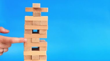 Bir kadın mavi parlak arka planda Jenga oynuyor. Aile tatili, tatiller, eğlence. Bir kadının eli, tahta oyuncak kulesinden bir blok ötede. Oyun