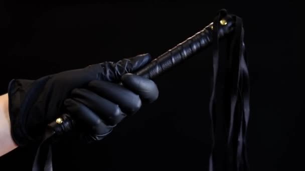 黒いラテックスグローブの手は 暗い背景に対する鞭を保持しています ブラックで隔離されたスパンキング用のレザーホイップ 親密さのためのセックスのおもちゃ 性奴隷制 — ストック動画