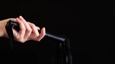 Bir kadının eli, koyu bir arka planda kamçıyı tutar. Siyah üzerine izole edilmiş kıçına şaplak atmak için deri kamçı. Samimiyet için seks oyuncağı. Cinsel kölelik