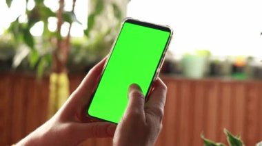 Bir kadının eli yeşil ekranlı bir telefonu tutuyor. Chroma anahtarı, akıllı telefon teknolojisi, bir kadının elindeki cep telefonu bir apartmanın arka planında. Cep telefonu tutan bir kadının yakın çekimi.