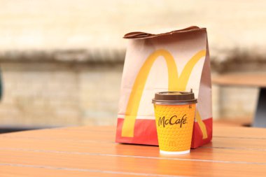 Kyiv, Ukrayna - 19 Mart 2024: Üzerinde McDonalds 'ın fast food restoranı logosu ve sokaktaki masada bir fincan kahve olan bir kağıt torba. Bulanık arka planda paket yemek torbası. Latte