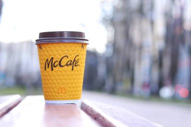 Kyiv, Ukrayna - 19 Mart 2024: Caddedeki bankta üzerinde McDonald 's fast food restoran logosu olan bir kağıt latte fincanı. Arka planda bulanık bir kahve. Tek kullanımlık sarı karton bardakta Latte.