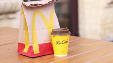 Kyiv, Ukrayna - 19 Mart 2024: Üzerinde McDonalds 'ın fast food restoranı logosu ve sokaktaki masada bir fincan kahve olan bir kağıt torba. Bulanık arka planda paket yemek torbası. Latte