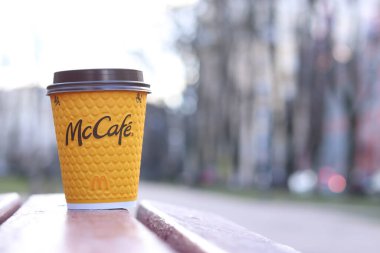 Kyiv, Ukrayna - 19 Mart 2024: Caddedeki bankta üzerinde McDonald 's fast food restoran logosu olan bir kağıt latte fincanı. Arka planda bulanık bir kahve. Tek kullanımlık sarı karton bardakta Latte.