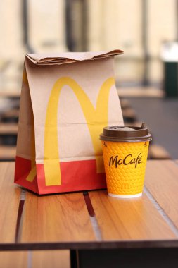 Kyiv, Ukrayna - 19 Mart 2024: Üzerinde McDonalds 'ın fast food restoranı logosu ve sokaktaki masada bir fincan kahve olan bir kağıt torba. Bulanık arka planda paket yemek torbası. Latte