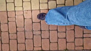 Sokakta yürürken insan ayağına yakın çekim. Bahar ya da yaz aylarında parkta yürüyüş. Yürüyüş. Mavi kotlu ve spor ayakkabılı kadın bacakları fayanslı bir sokakta yürüyor, birinci şahıs manzaralı.