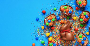 Çikolata tavşanı ve şeker, mavi arka planda renkli şeker parçacıkları, üst manzara. Paskalya kompozisyonu. Paskalya çikolatalı tavşanı. Şenlikli tatlı ikram. Çikolata heykeli. Tatlılar