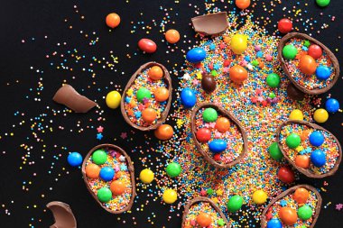 Şekerli çikolata yumurtaları, siyah arka planda şeker renkli şekerler, üst manzara. Paskalya kompozisyonu. Masanın üzerinde yarım çikolatalı yumurta. Şenlikli tatlı ikram. Tatlılar