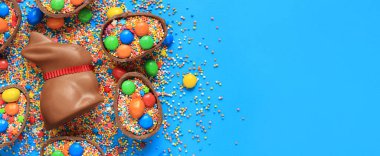 Çikolata tavşanı ve şeker, mavi arka planda renkli şeker parçacıkları, üst manzara. Paskalya kompozisyonu. Paskalya çikolatalı tavşanı. Şenlikli tatlı ikram. Çikolata heykeli. Tatlılar