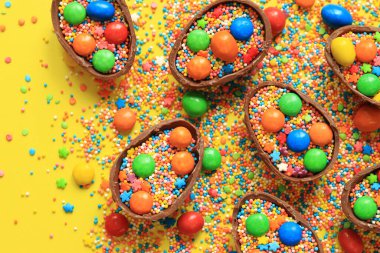 Çikolata yumurtaları ve şeker renkli şekerler sarı arka planda, üst manzara. Paskalya kompozisyonu. Masanın üzerinde yarım çikolatalı yumurta. Şenlikli tatlı ikram. Tatlılar