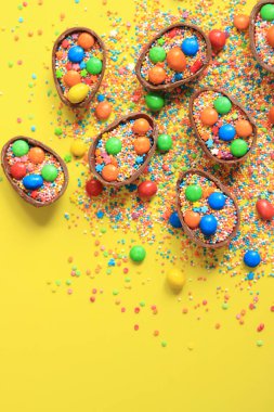 Çikolata yumurtaları ve şeker renkli şekerler sarı arka planda, üst manzara. Paskalya kompozisyonu. Masanın üzerinde yarım çikolatalı yumurta. Şenlikli tatlı ikram. Tatlılar