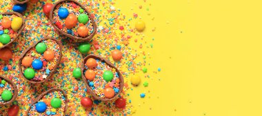 Çikolata yumurtaları ve şeker renkli şekerler sarı arka planda, üst manzara. Paskalya kompozisyonu. Masanın üzerinde yarım çikolatalı yumurta. Şenlikli tatlı ikram. Tatlılar