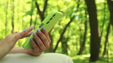 Elleri cep telefonuyla yakın plan. İnternette sörf yap. Parkta telefonu olan bir kız, Summer. Akıllı telefonlu kadın ellerinin yakın çekimi. Modern teknoloji aygıtı kavramları. Kadın ve telefon