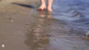 Sahilde çıplak erkek ayakları. Yazın nehrin yanında yürü. Suya yakın kumda duran insan ayaklarının yakın çekimi. Kumsal. Yazın nehirde dinlen. Adam ayaklarını nehir suyunda ıslatıyor.